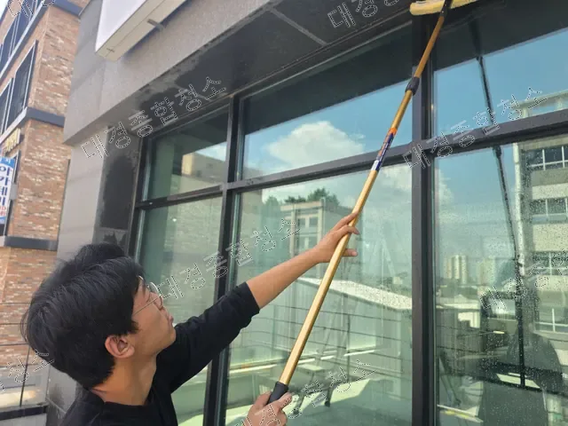 장대 도구로 외벽 유리창을 청소하는 안경 쓴 작업자